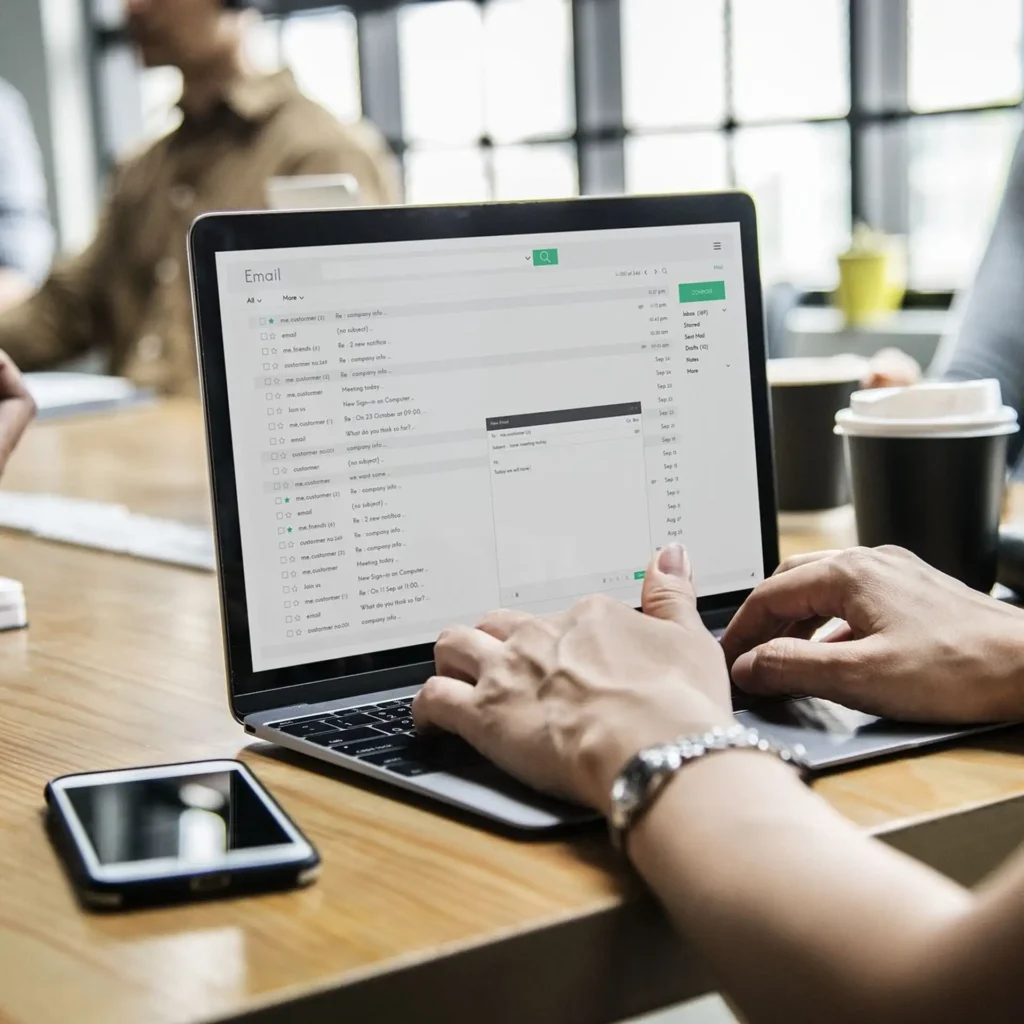 Person typing on a laptop displaying an email inbox interface while sitting at a wooden office table